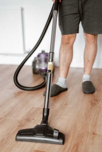 A yellow canister vacuum cleaner with a hose attachment is positioned on a decorative rug in a living room area, coming from a round metal pipe. The room features wooden flooring and a black metal TV 